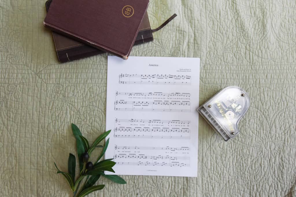 Sheet music to "America". The music is surrounded by a model piano, a bible, and a small leafy plant.