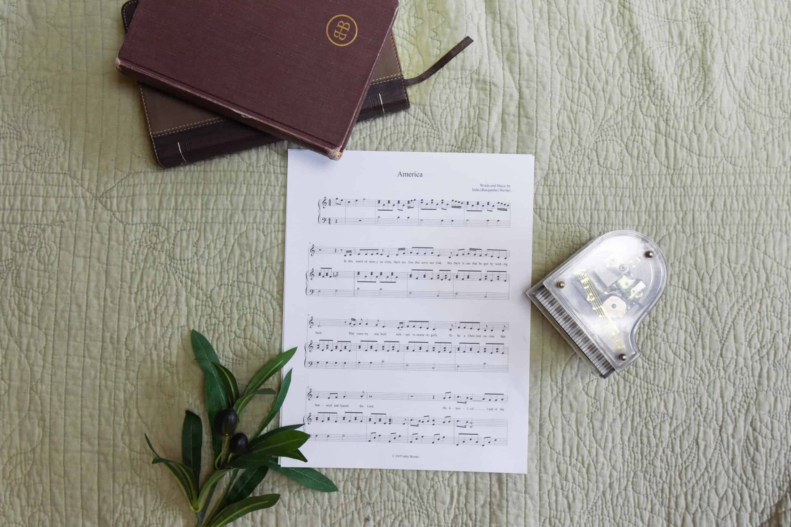 Sheet music to "America". The music is surrounded by a model piano, a bible, and a small leafy plant.