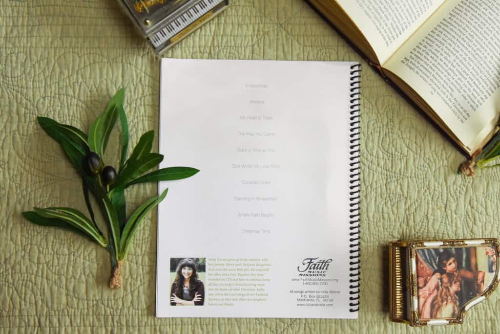 Sheet music back cover of "Where Faith Begins" sitting on a table with a model piano, bible, and plant.