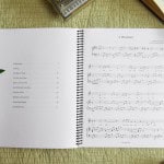 Sheet music book "Where Faith Begins" laying open with table of contents showing. On the table is a bible, model piano, and a green plant.