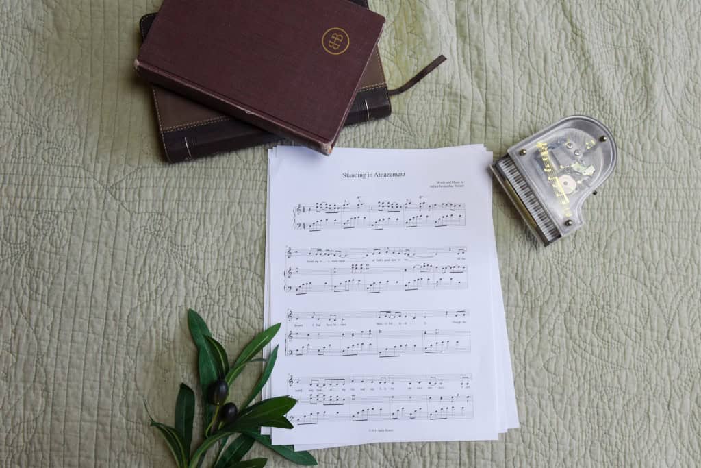 Sheet music to "Standing in Amazement". The music is surrounded by a model piano, a bible, and a small leafy plant.